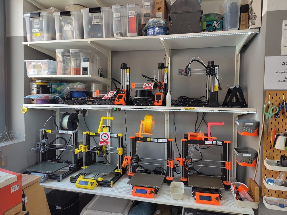 Estante de um hackerspace com várias impressoras 3D Prusa lado a lado e caixas de filamento nas prateleiras de cima.