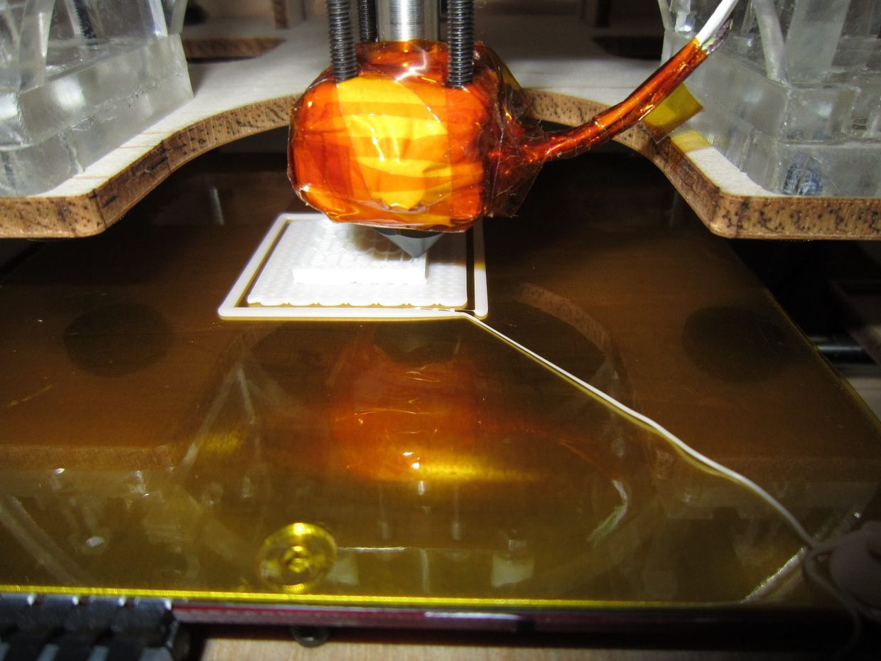 Close do cabeçote de uma impressora 3D depositando filamento branco sobre a mesa, com as primeiras camadas de uma peça já formadas.