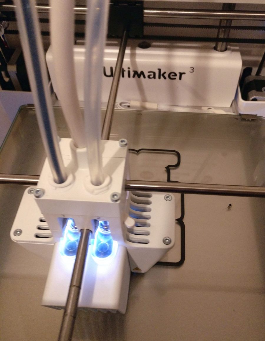 Cabeçote duplo branco de uma impressora Ultimaker 3 em funcionamento, com dois tubos de filamento e luzes azuis sobre a mesa de vidro.