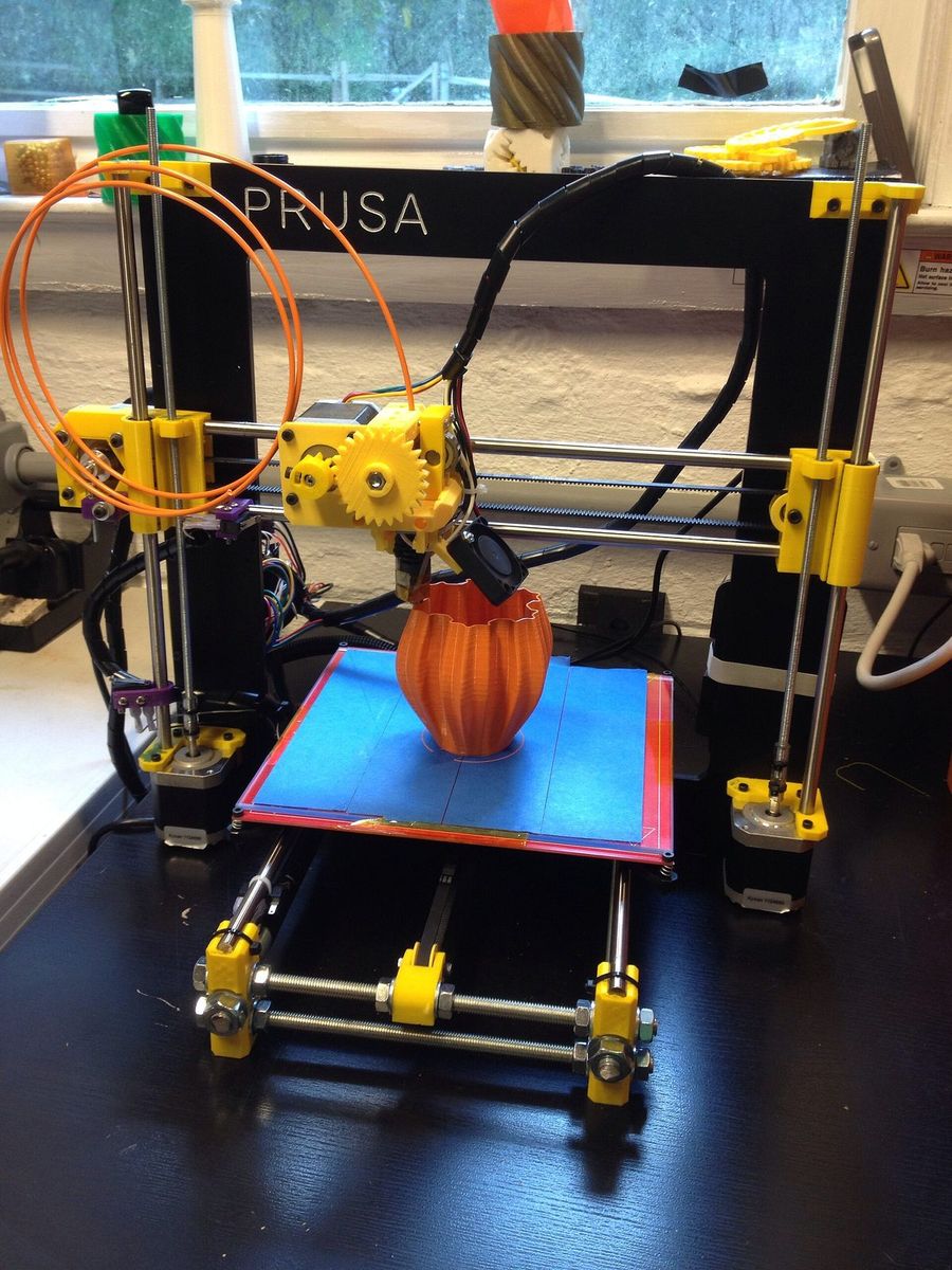 Impressora 3D Prusa i3 de estrutura aberta imprimindo um vaso laranja sobre a mesa coberta com fita azul.