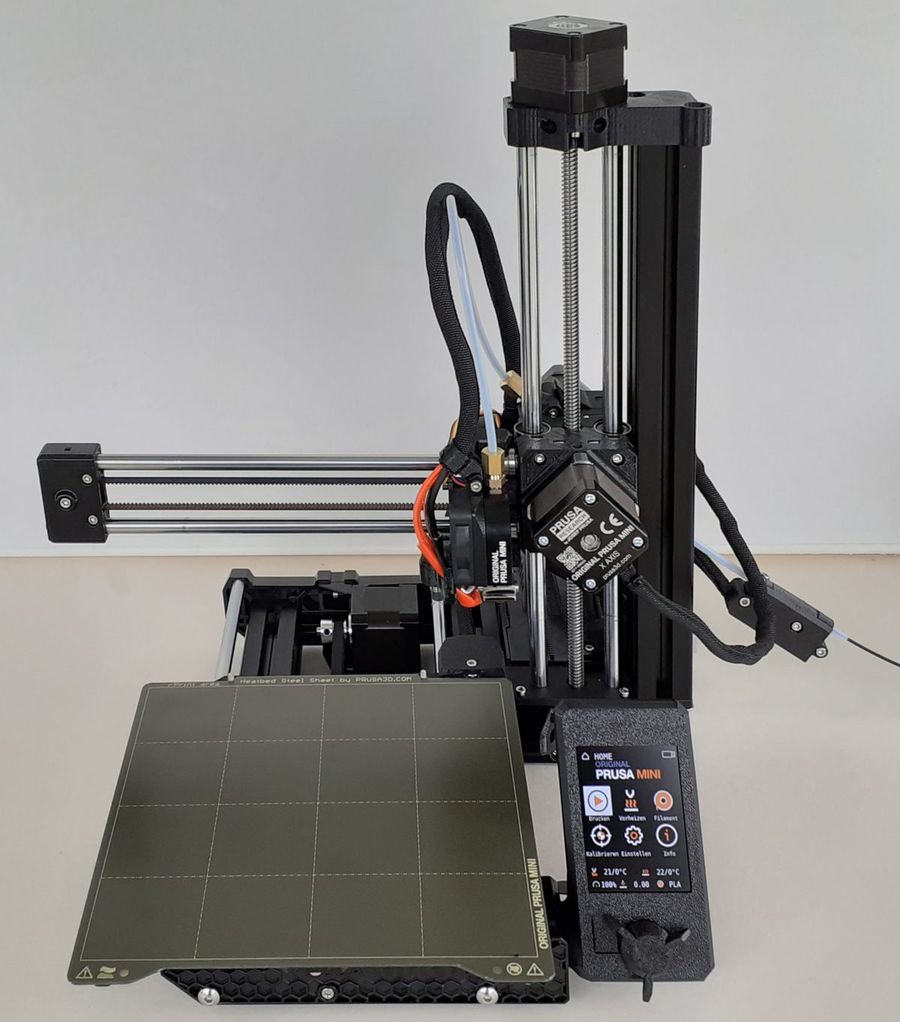 Impressora 3D Prusa Mini+ de frente, com a placa de aço da mesa à mostra e a tela de controle ao lado.